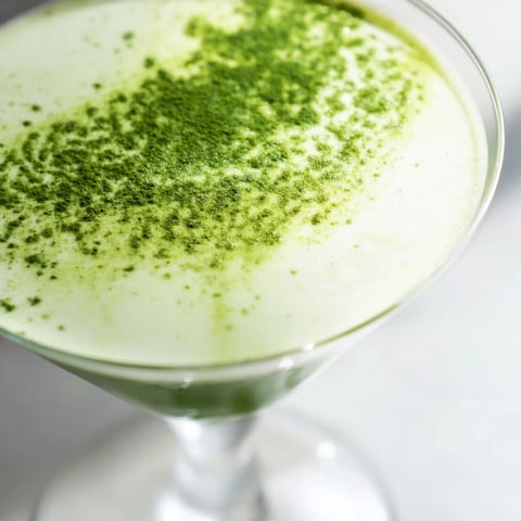 A chilled Matcha Martini cocktail garnished with matcha powder sits in a classic stemmed glass on a wooden surface.  